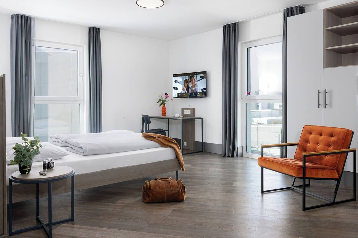A modern hotel room with a bed, a desk, a chair, and a wardrobe.