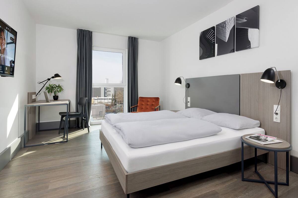 A modern hotel room with a large bed, a desk with a laptop, a chair, a small table with a magazine, and a view of the city through the window.