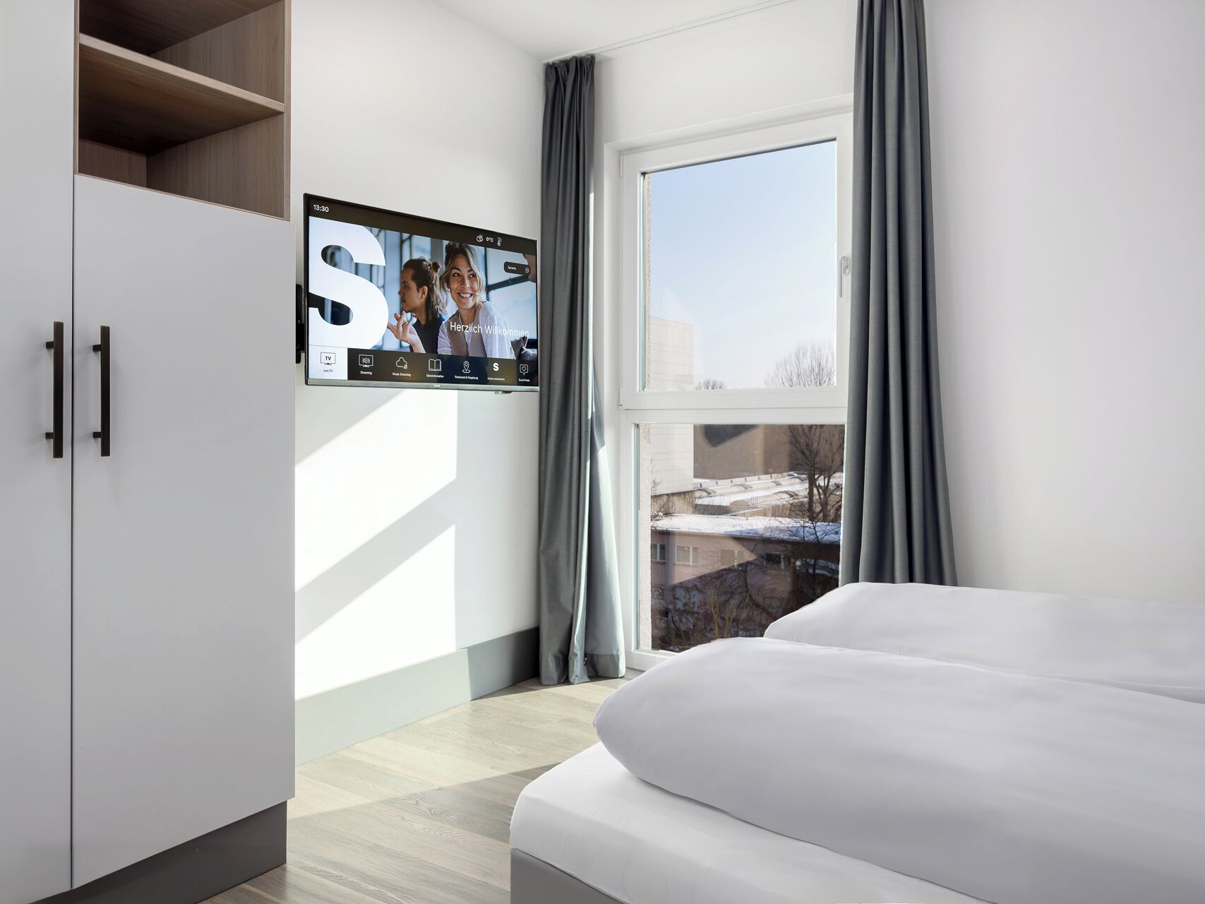 A modern hotel room with a large window, a flat-screen TV mounted on the wall, and a bed with white bedding.