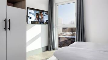 A modern hotel room with a large window, a flat-screen TV mounted on the wall, and a bed with white bedding.