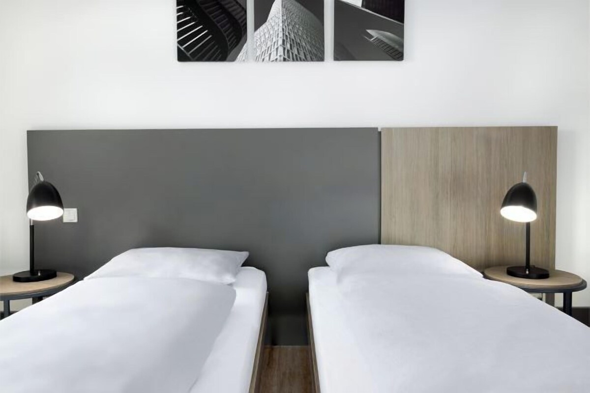 Habitación de hotel con dos camas individuales, cabecero gris, panel decorativo de madera y dos lámparas de noche.