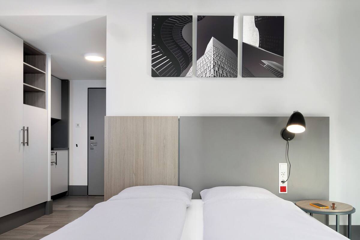 A modern hotel room with a large bed, a desk with a lamp, and a view of a building through the window.