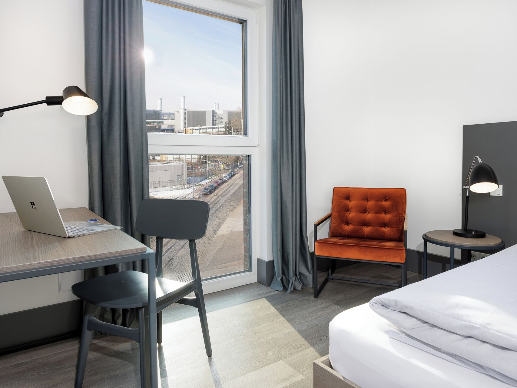 Une chambre d’hôtel avec un lit, un bureau avec un ordinateur portable, une chaise, une petite table avec une lampe, et une vue sur une voie ferrée et des immeubles.