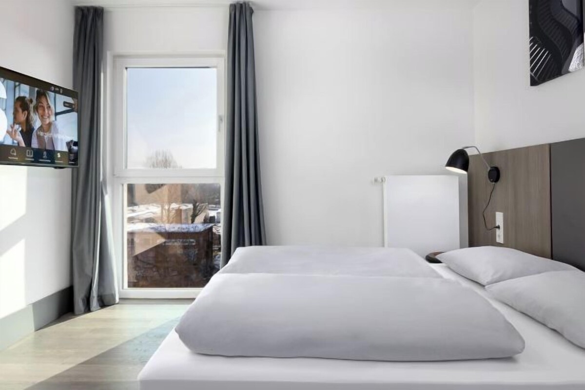 Habitación de hotel con cama, televisión, ventana con cortinas y vistas al exterior.