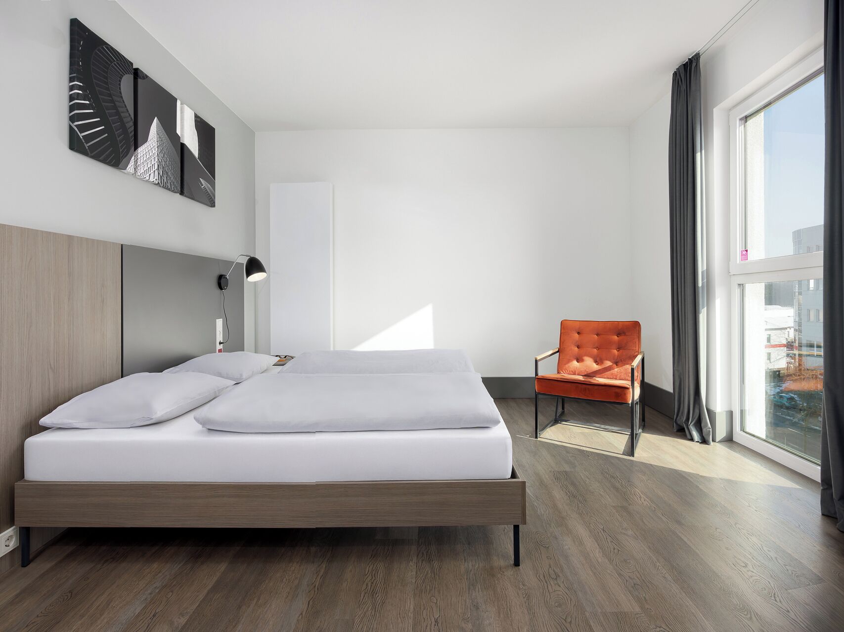 A modern bedroom with a wooden bed, a single chair, a large window, and a city view.