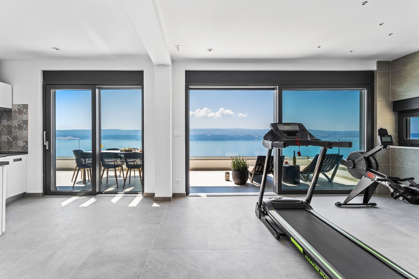 A modern living room with a treadmill, a dining area with a view of the sea, and a large sliding glass door leading to a balcony.