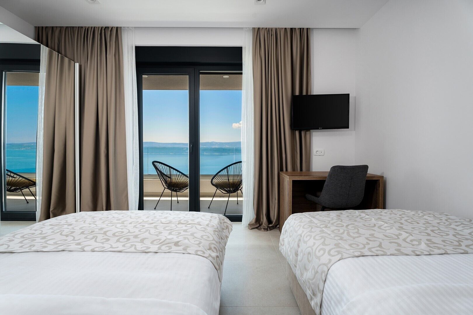 Habitación de hotel con dos camas, un escritorio, un televisor y un balcón con sillas.