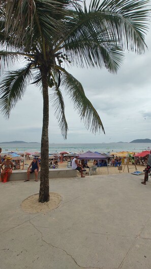 A beach scene with palm trees, beachgoers, and umbrellas. - Lindo APT inteiro mt perto da praia Peró (Cabo Frio)