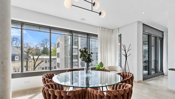 Un'area pranzo moderna con un tavolo in vetro circondato da confortevoli poltrone marroni, ampie finestre con vista sulla città e un arredamento minimalista con un vaso di piante.