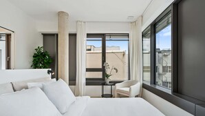 Una camera d'albergo moderna con un letto grande, un tavolino con una pianta in vaso, una sedia e vista sulla città attraverso ampie finestre.