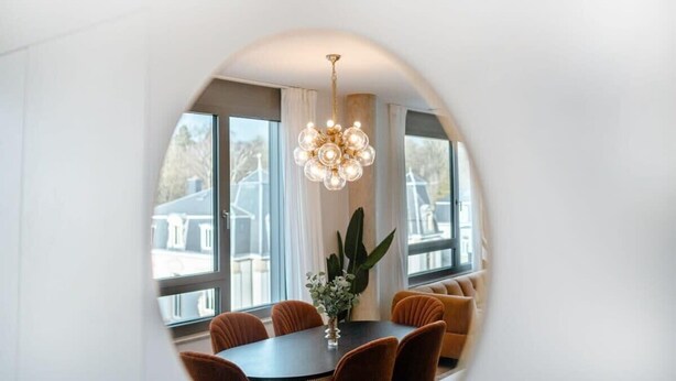 Un coin repas avec une table ronde, des chaises orange, un lustre et de grandes fenêtres avec des rideaux.