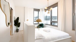 Une chambre moderne avec un grand lit, une table de chevet, une plante en pot et une vue sur la ville par la fenêtre.