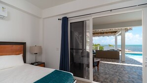 Een hotelkamer met een bed, nachtkastje, lamp en uitzicht op een zwembad en zee door een schuifdeur.