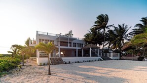Een modern strandhuis met een tweelaags gebouw, een overdekt terras en een zandstrand op de voorgrond.