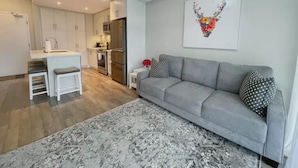 Een moderne woonkamer met een grijze bank, een kamerkleed met patroon en een keuken op de achtergrond.