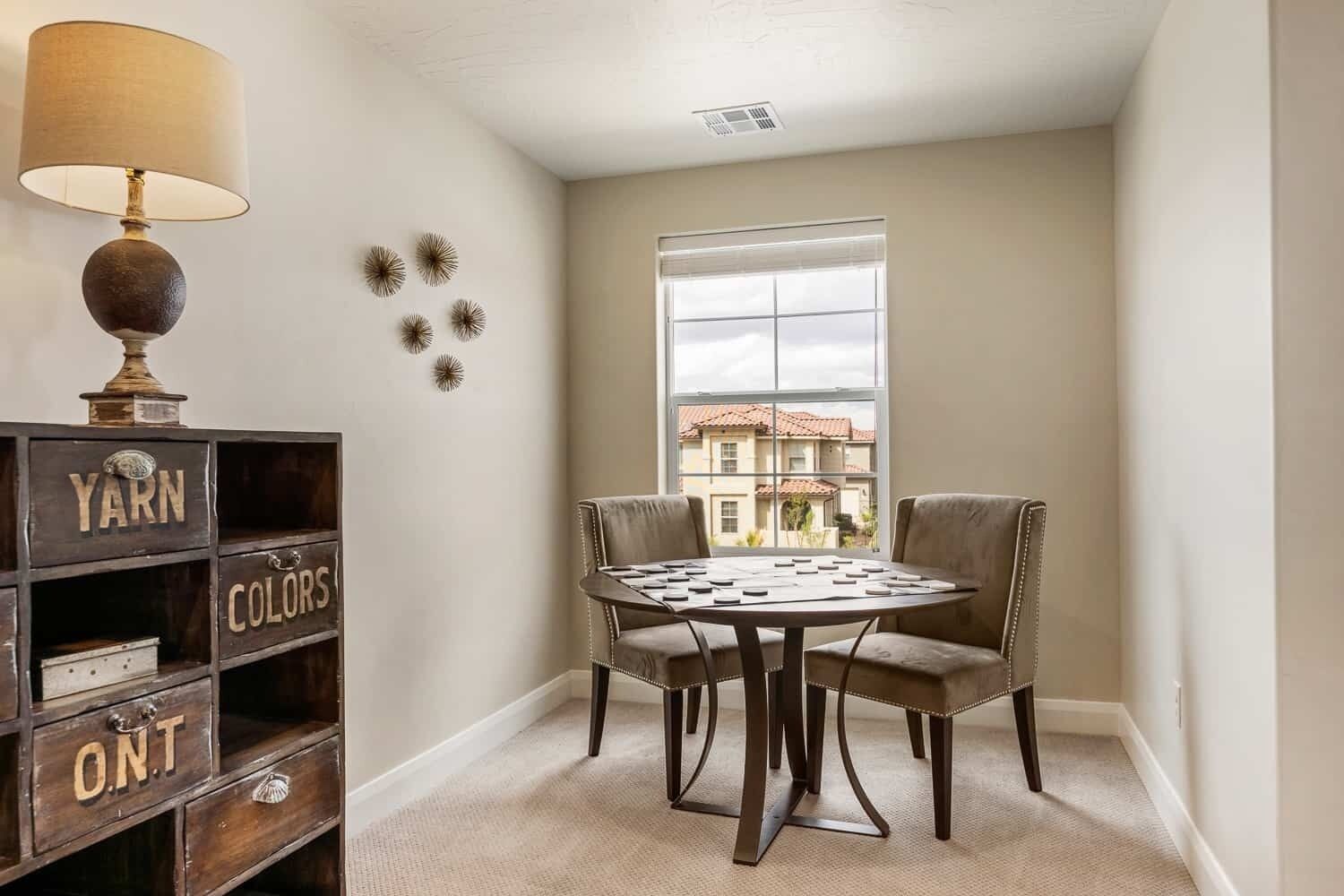 A corner room with a table and chairs, a lamp, a shelf with storage boxes, and a window with a view of buildings.