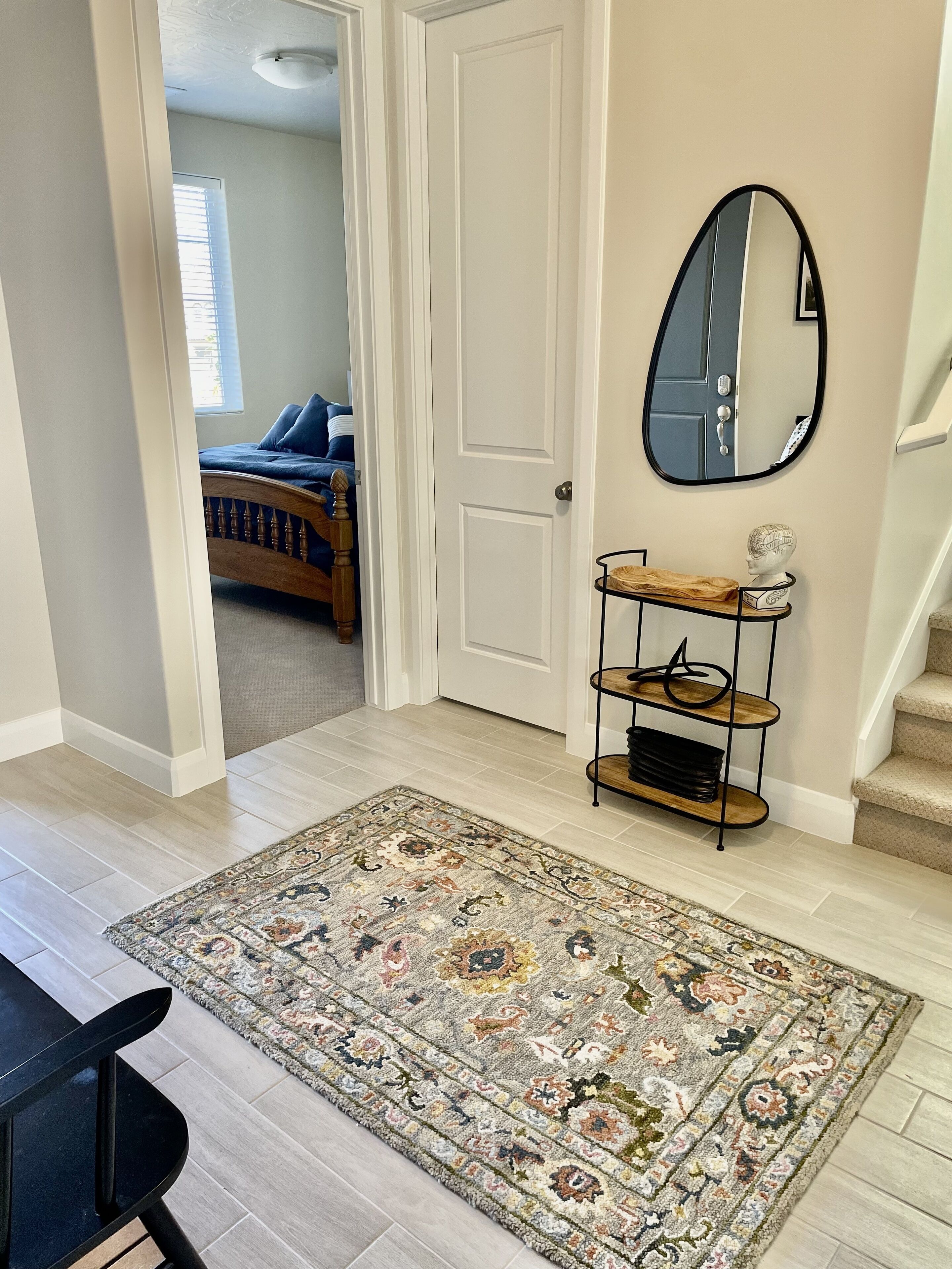 A hallway with a patterned rug, a wooden bench, a metal shelf with decorative items, and a door leading to a bedroom with a bed and blue pillows.