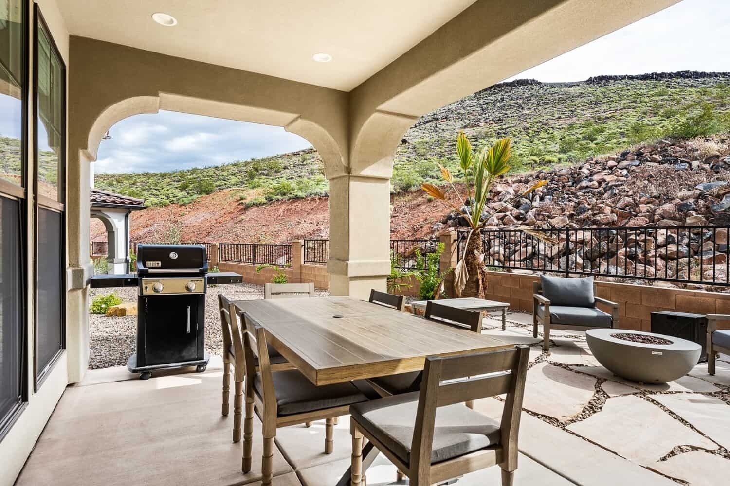 Een terras buiten met een houten tafel en stoelen, een barbecue en een zithoek met een vuurkorf.