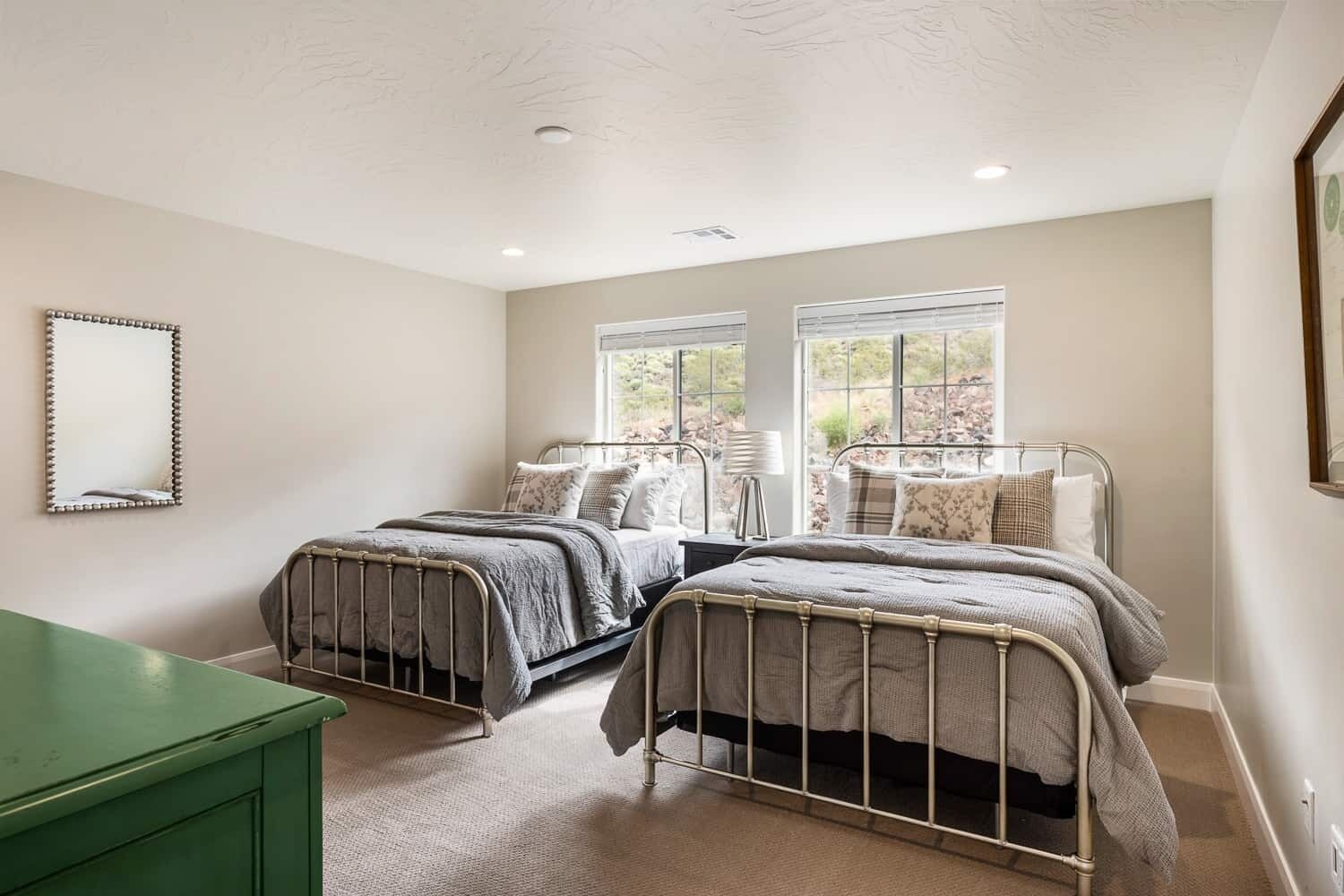 A bedroom with two metal-framed beds, a green dresser, and a large window providing a view of the outdoors.
