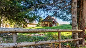 A rustic cabin with a wooden deck and picnic table, surrounded by trees and a grassy area.