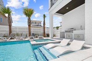 Un área de piscina con sillones de descanso, una parrilla y un muro de ladrillo blanco.
