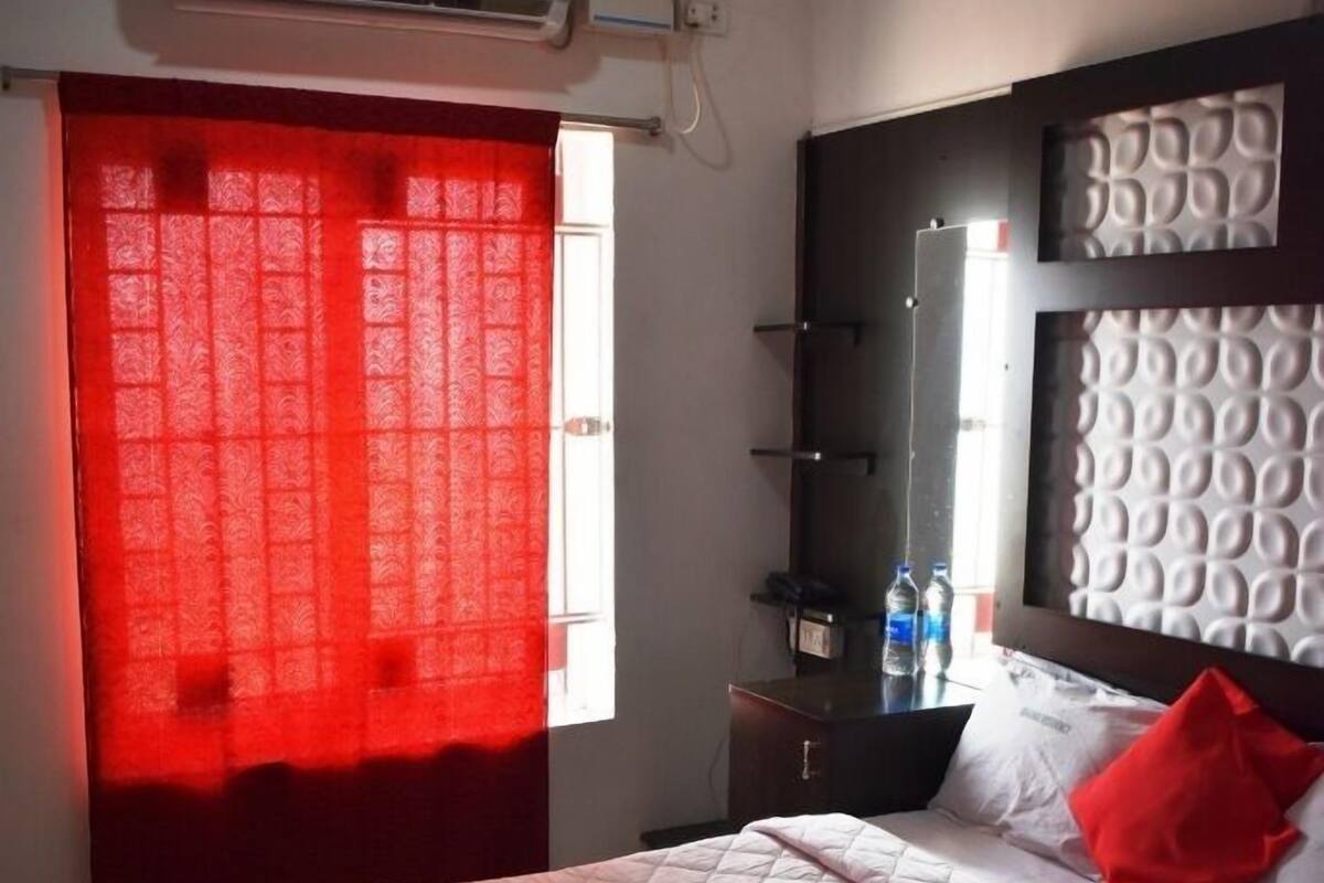 Hotelkamer met een bed, een plafondventilator, een airconditioner, een raam met rode gordijnen, een nachtkastje met waterflesjes en een hoofdbord met een structuur.