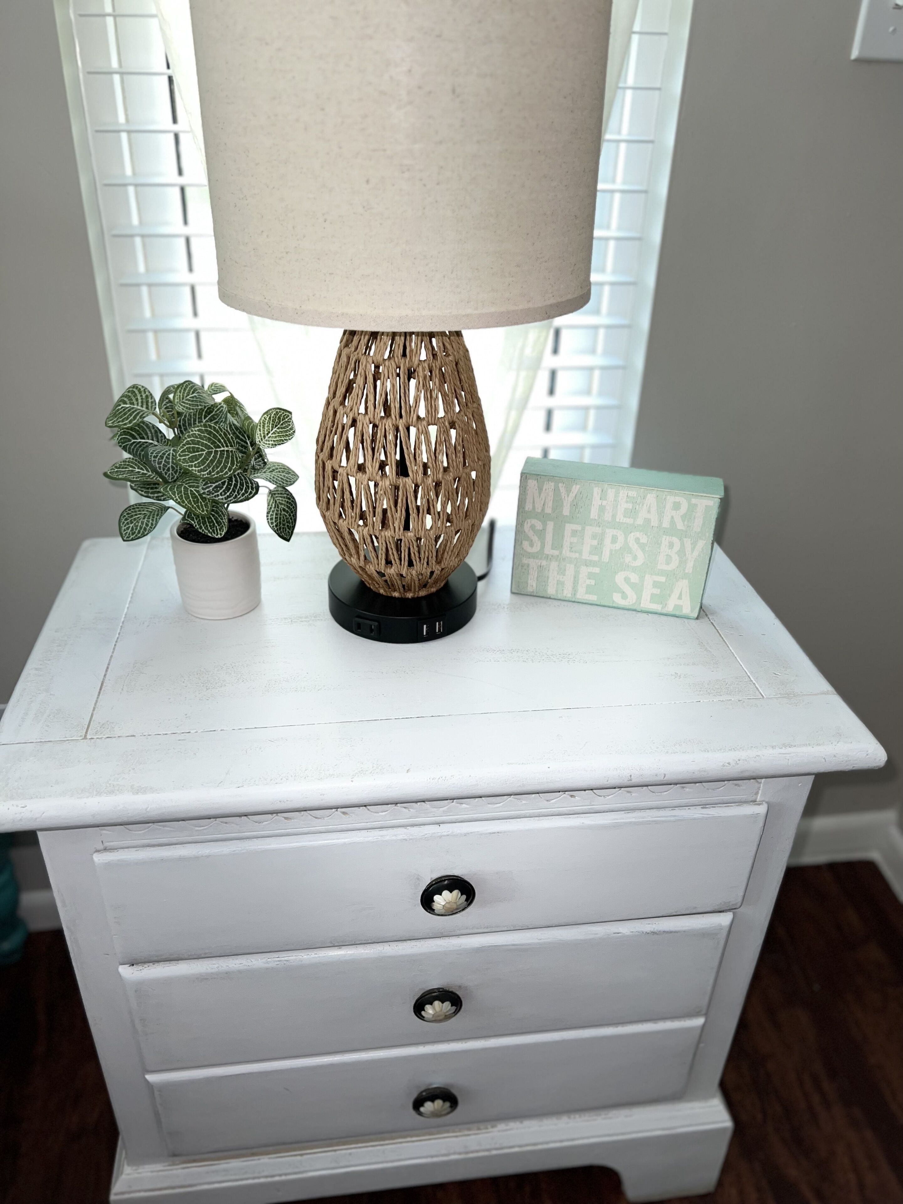 A white nightstand with a woven lamp, a small potted plant, and a sign that reads "MY HEART SLEEPS BY THE SEA".