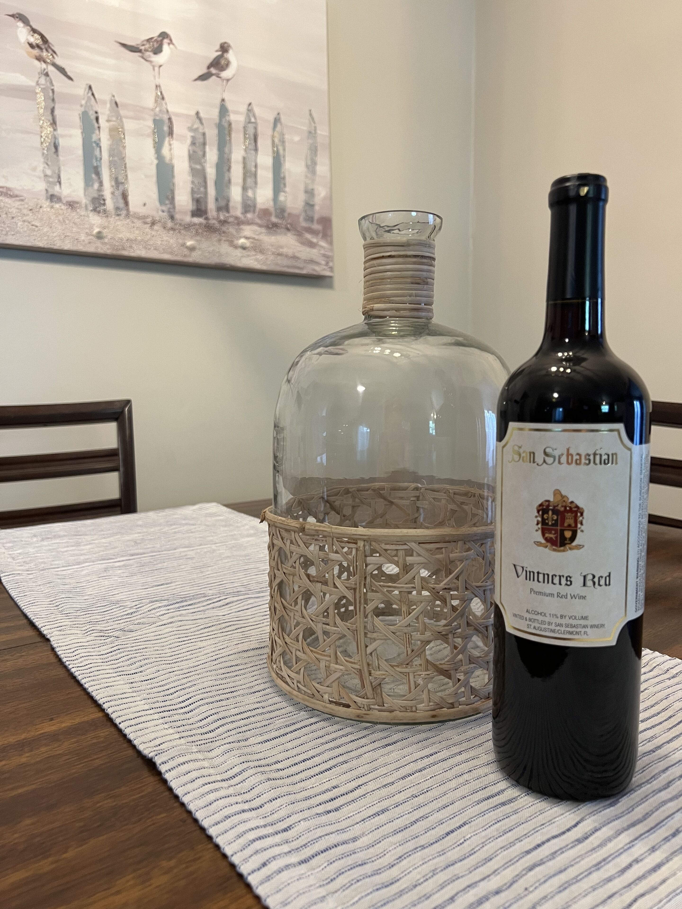 A bottle of Vintners Red wine and a decorative glass decanter on a table with a woven placemat.