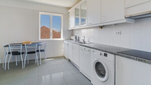 A modern kitchen with white cabinets, a built-in washing machine, and a dining area with a table and chairs. - Bela São Tiago I (Funchal)