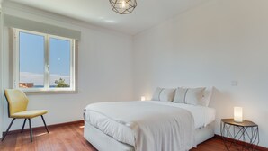 A bedroom with a large bed, a chair, a bedside table with a lamp, and a window with a view of the sky. - Bela São Tiago I (Funchal)