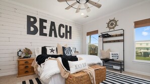 Une chambre avec un grand panneau « PLAGE » accroché au mur, un lit recouvert d’un tapis à rayures, une table de chevet en bois, un panier en osier et un porte-manteau décoré d’un volant de navire.