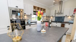 A modern kitchen with white cabinets, a built-in oven, a microwave, and a stainless steel range hood. There is a vase with tulips on the counter, a bottle of wine, and a candle holder. - 48 Crosswinds (Bembridge)