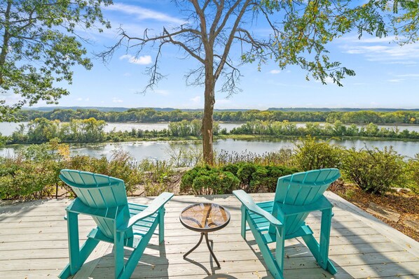 A wooden deck with two turquoise chairs facing a lake, surrounded by greenery and a clear sky.