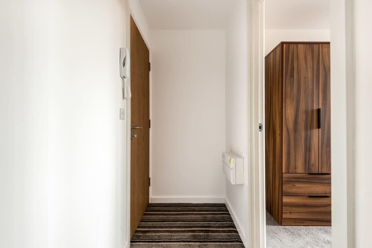 Een smalle gang met een houten deur aan de linkerkant, een witte muur en een houten garderobe aan de rechterkant.