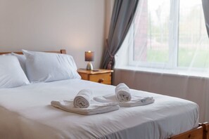 A neatly made bed with white linens and towels, a bedside lamp, and a window with curtains.