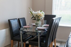 A dining table with four chairs, a vase with flowers, and a window in the background.