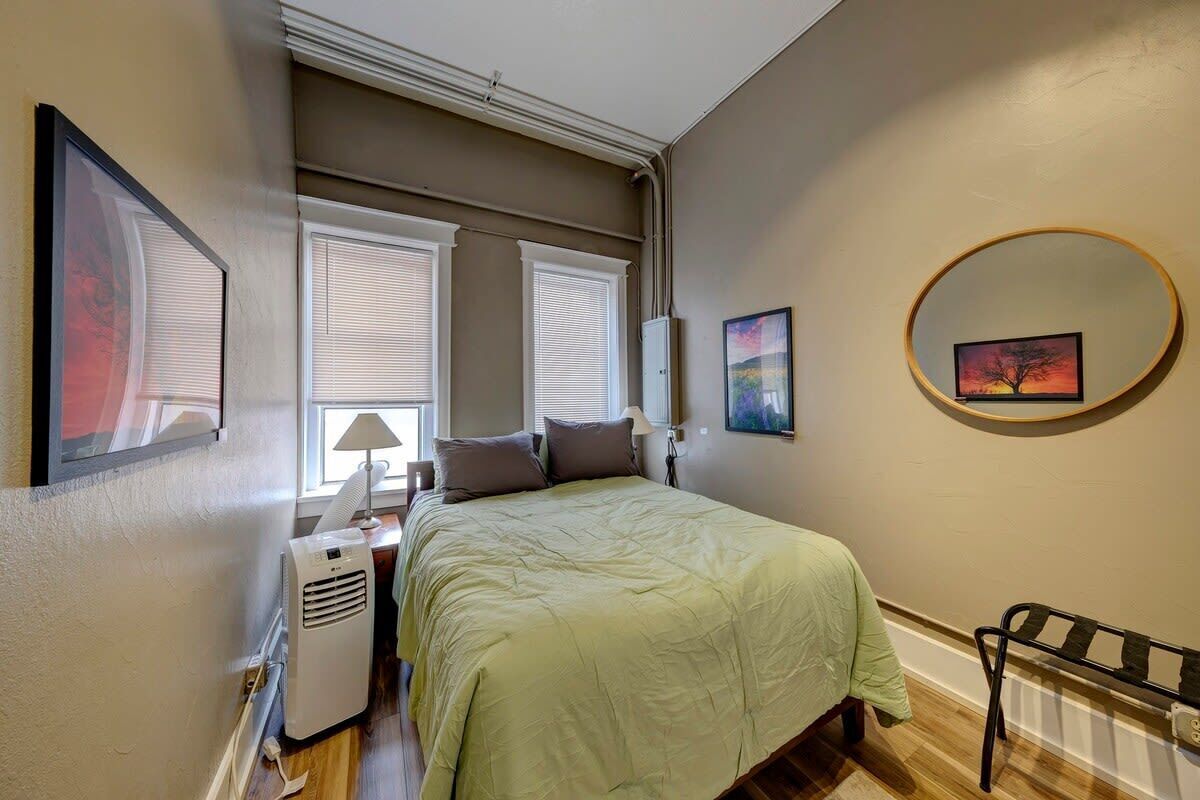 A bedroom with a bed, two windows with blinds, a heater, a lamp, and a wall-mounted mirror.