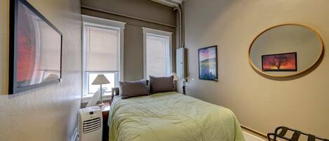 A bedroom with a bed, two windows with blinds, a heater, a lamp, and a wall-mounted mirror.