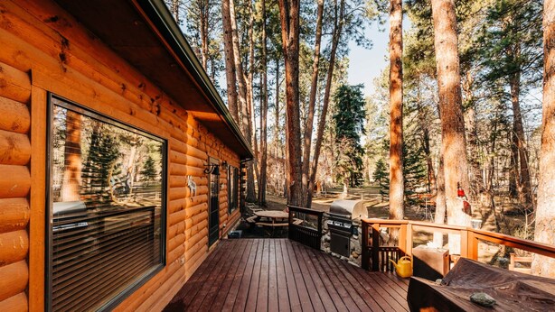 Eine Holzterrasse mit Holztisch und -stühlen, einem Grill und einem großen Fenster mit Blick auf einen Wald.