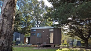 A small, two-story cabin with a metal roof, surrounded by trees and a grassy yard. - Tiny House 13 at Grampians' Edge by Tiny Away (Dadswells Bridge)