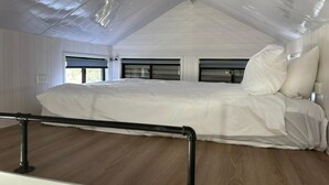 A bedroom with a sloped ceiling, a large bed, and a window with blinds. - Tiny House 13 at Grampians' Edge by Tiny Away (Dadswells Bridge)