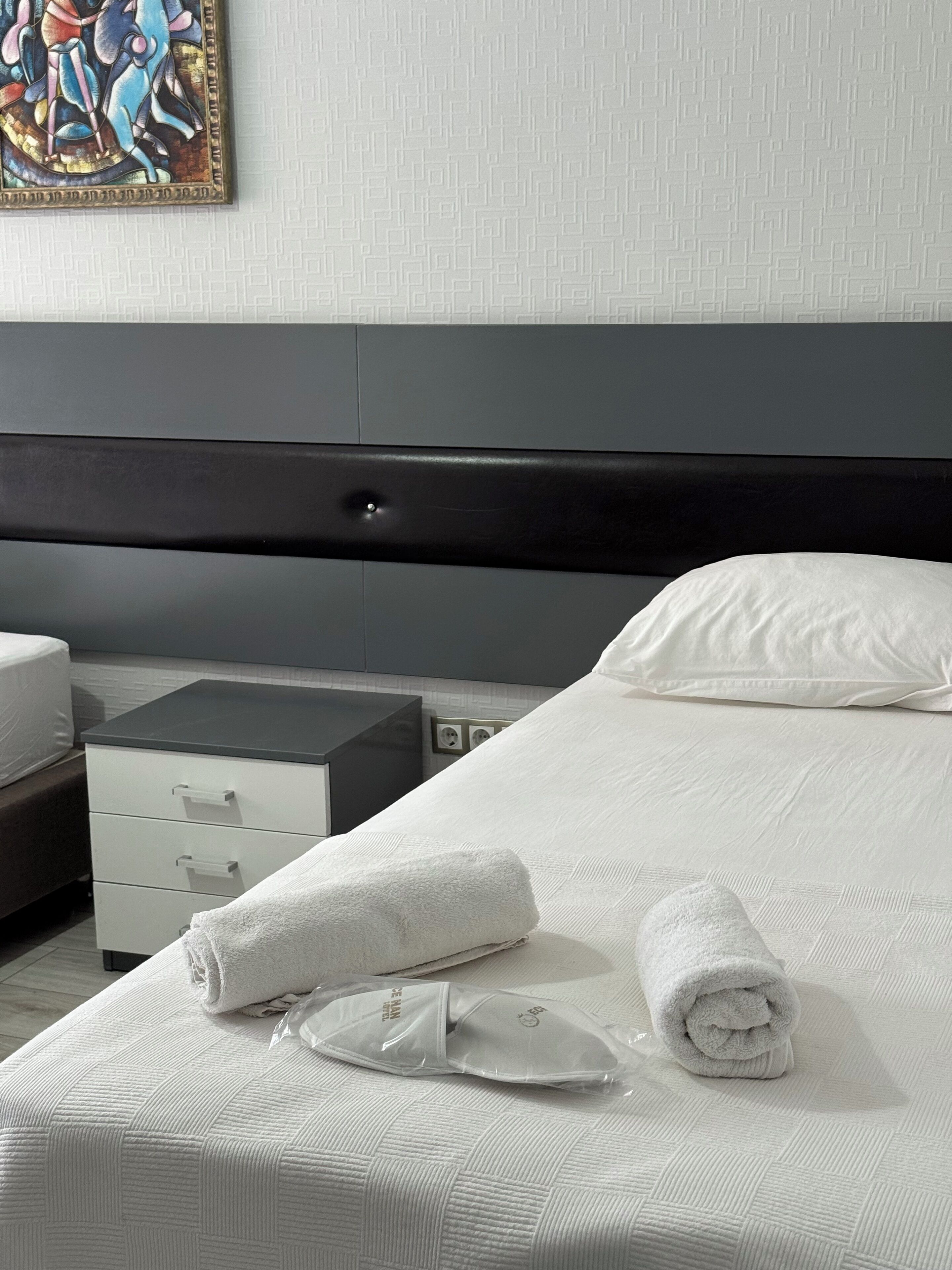 Una cama con ropa blanca de cama bien tendida, un cabecero gris y una almohada blanca. Hay dos toallas enrolladas sobre la cama.