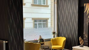 Una habitación con vistas a un edificio a través de una ventana, una silla amarilla, una mesita redonda y un armario blanco.