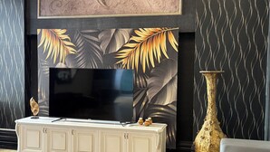 Una sala moderna con un televisor de pantalla plana grande montado en una pared con un estampado de hojas tropicales. Hay una consola de mesa color crema con objetos decorativos frente al televisor.