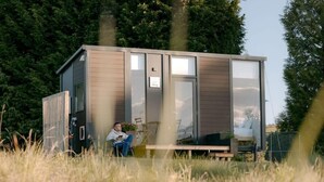 Eine moderne, kompakte Hütte mit Glastür, einer kleinen Terrasse und einer Person, die draußen auf einer Bank sitzt.
