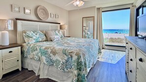 A bedroom with a bed, bedside tables, a ceiling fan, a TV, and a view of the beach.