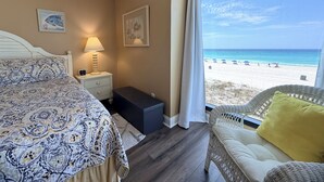 A bedroom with a bed, bedside table, lamp, and a view of the beach.