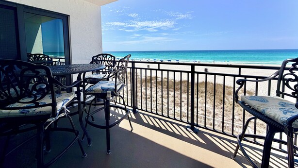A balcony with a view of the beach, a table, and chairs.