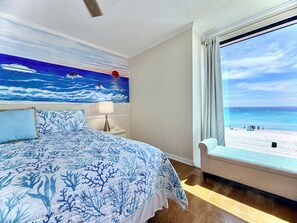 Un dormitorio con cama, mesita de noche, lámpara, un cuadro con escena de playa y ventana con vista al mar.