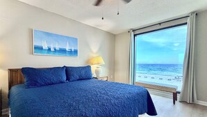 Une chambre avec un grand lit, un tableau représentant des voiliers, une table de chevet, une lampe et une vue sur la plage.
